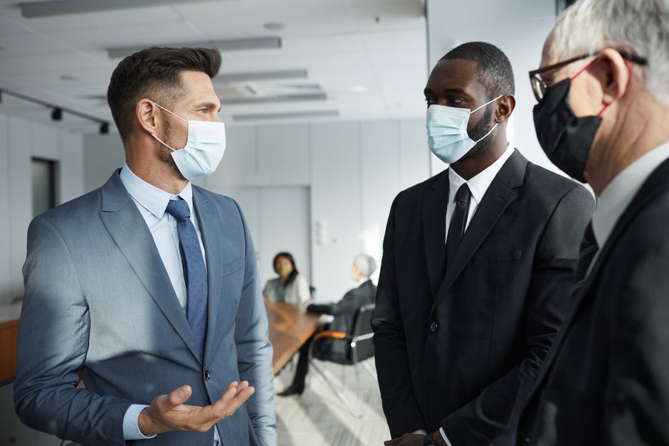 Businessmen wearing masks in a conference room discuss during a pandemic meeting. Suits and formal settings emphasize professionalism.