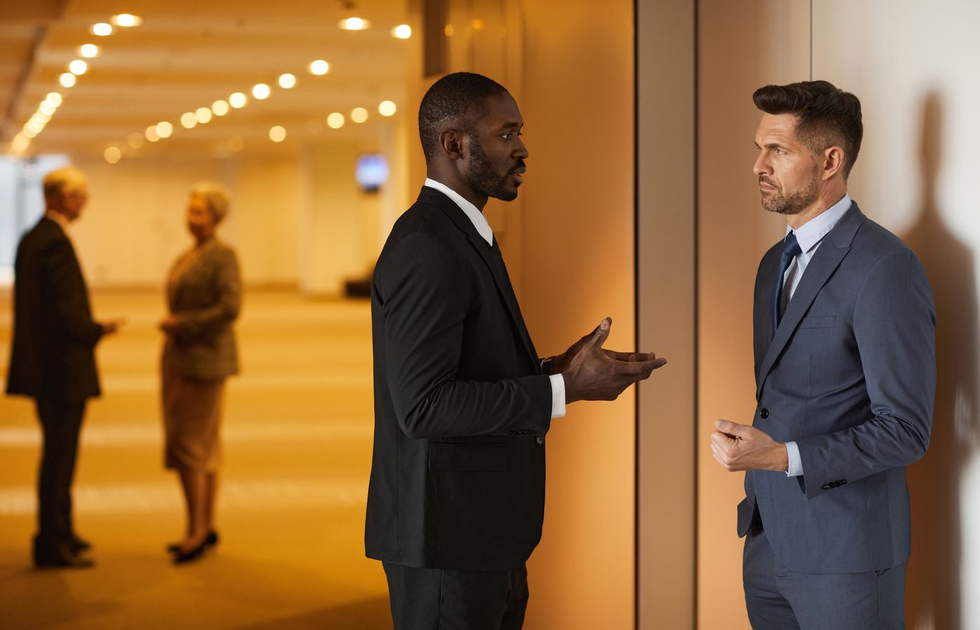 Two men in suits having a discussion in a modern hallway setting.