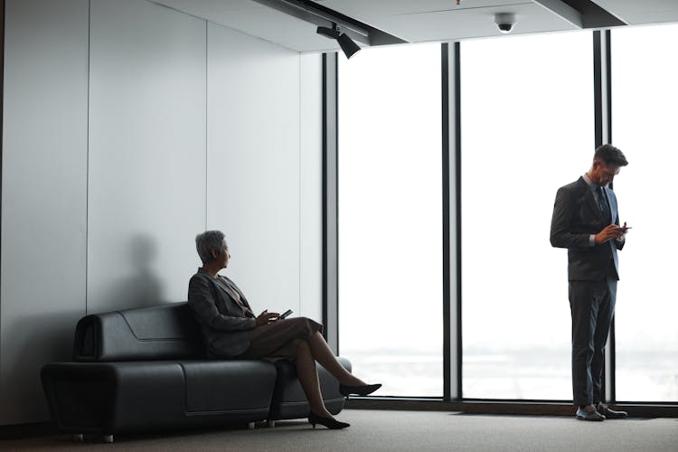 A Woman In Gray Blazer Sitting On The Couch Near The Man Standing Beside The Window