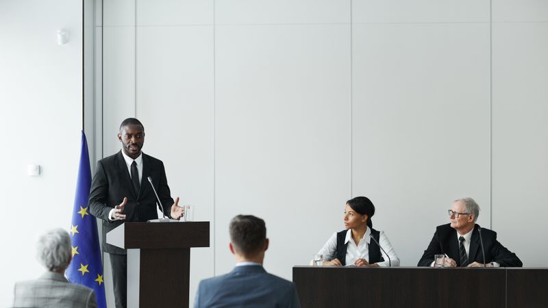Business meeting featuring diverse professionals discussing important topics with a speaker at the podium indoors.