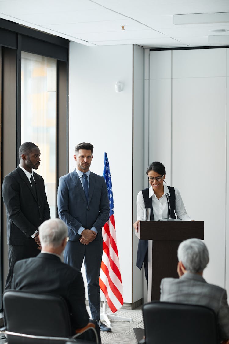 Men In Black And Gray Suit Standing Beside The Woman Talking At The Podium