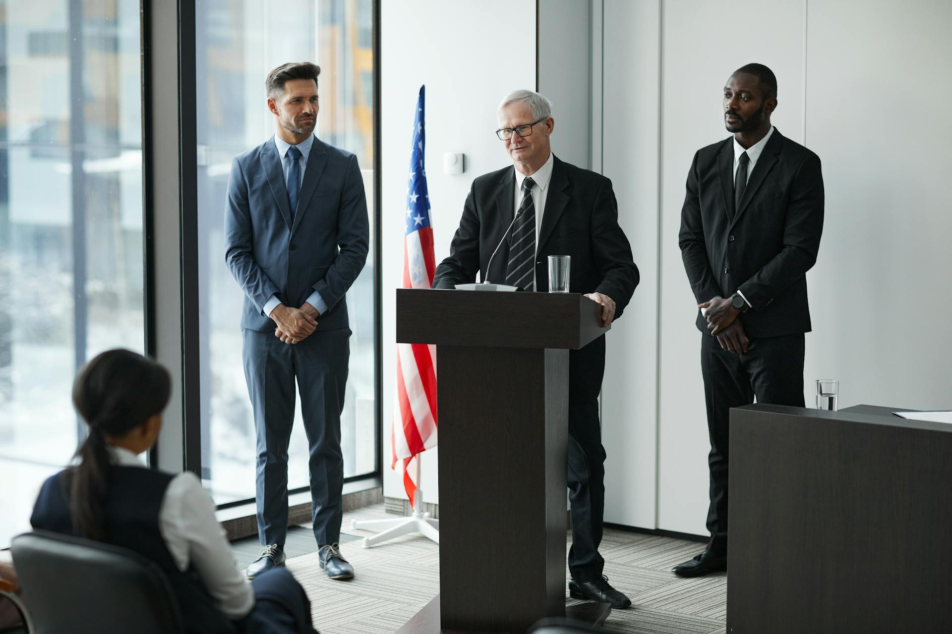 Officials delivering a political speech in a modern conference room with an American flag.