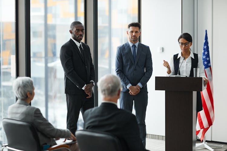 Woman On A Podium With Microphone Beside Men In Suits