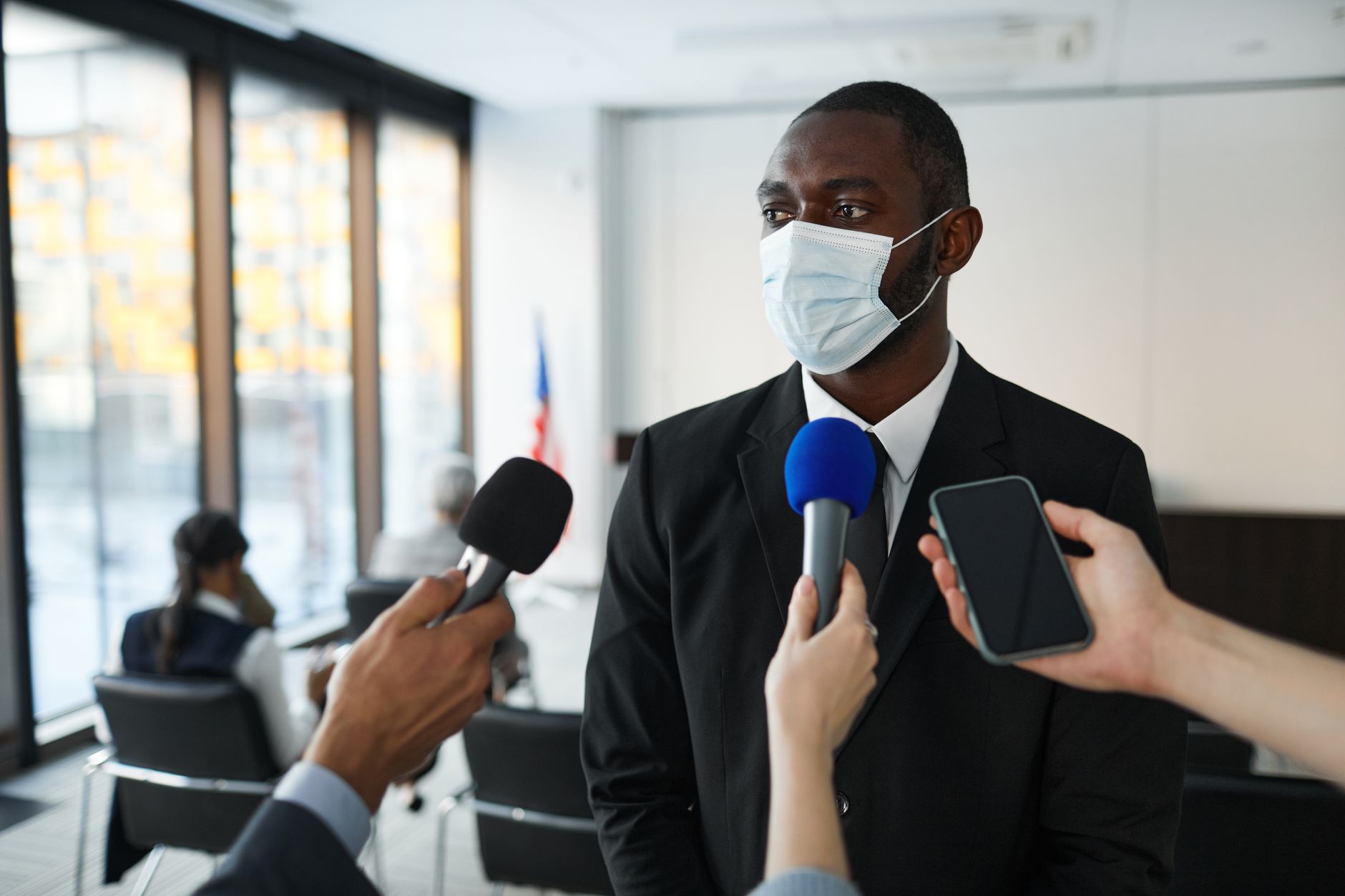 African American businessman in formal attire speaks to media while wearing a face mask in a conference room setting.