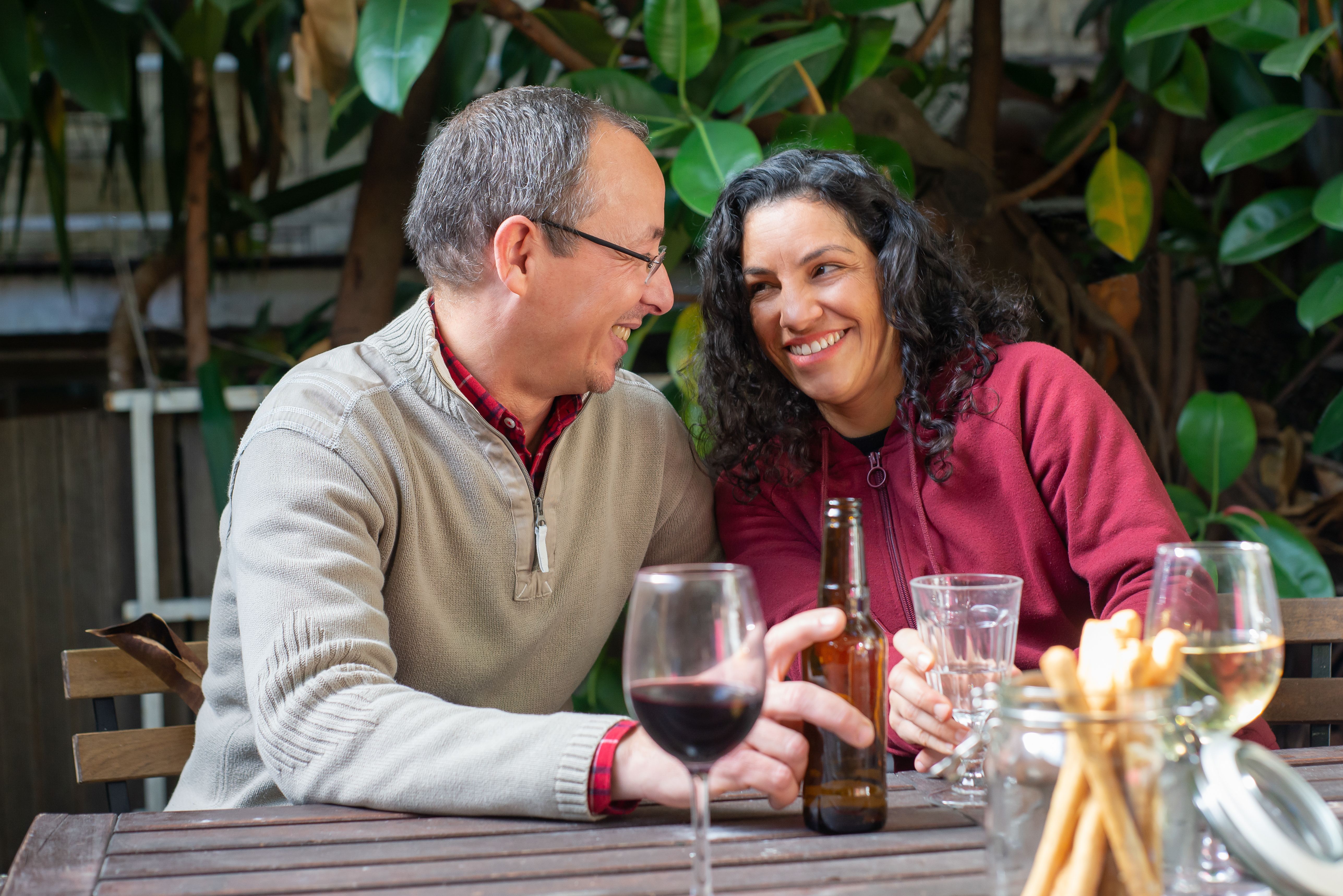 A cheerful couple sitting at a table, sharing smiles over wine and beer in an outdoor backyard.