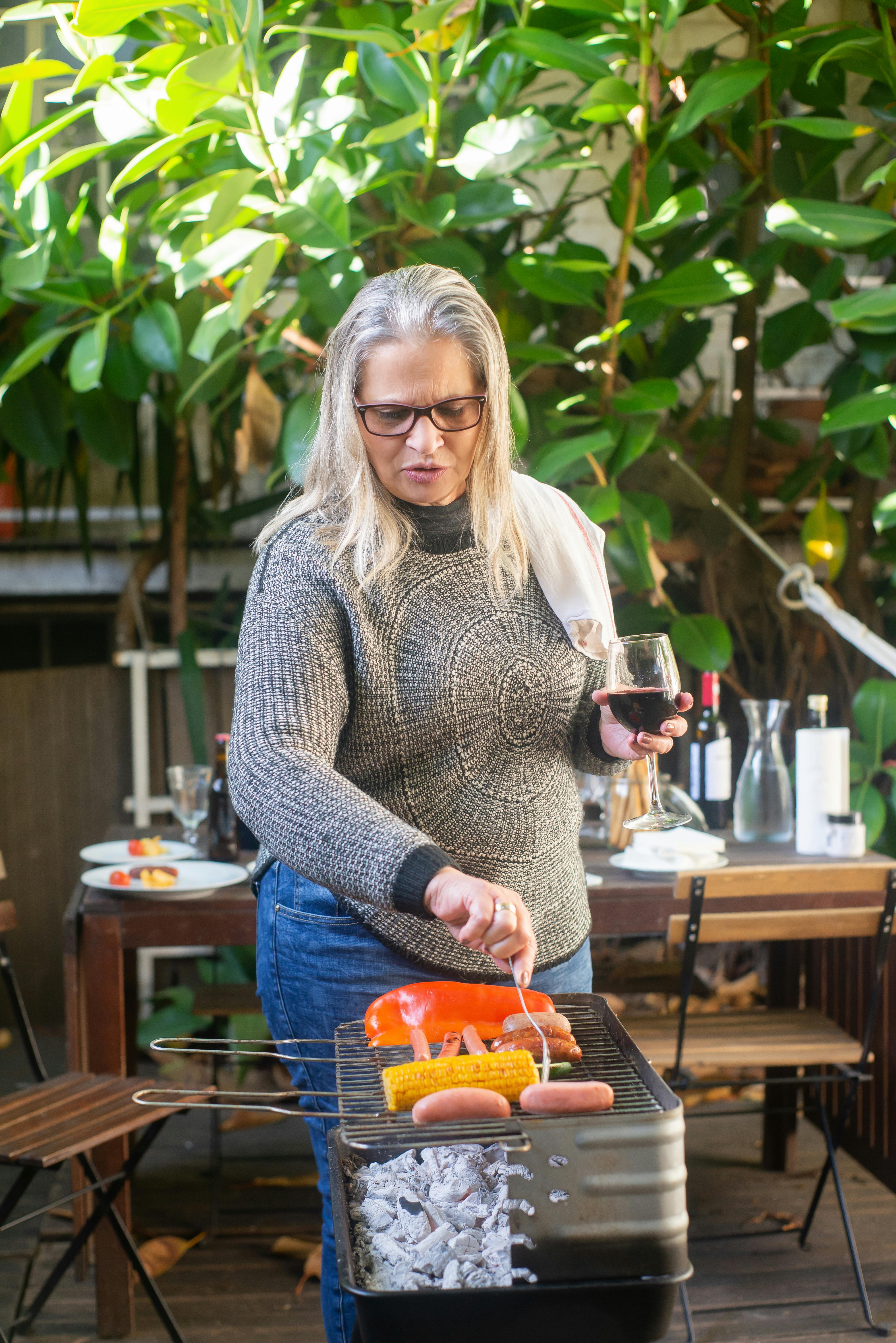 Senior woman enjoying outdoor barbecue with sausages and vegetables while holding wine in Portugal.