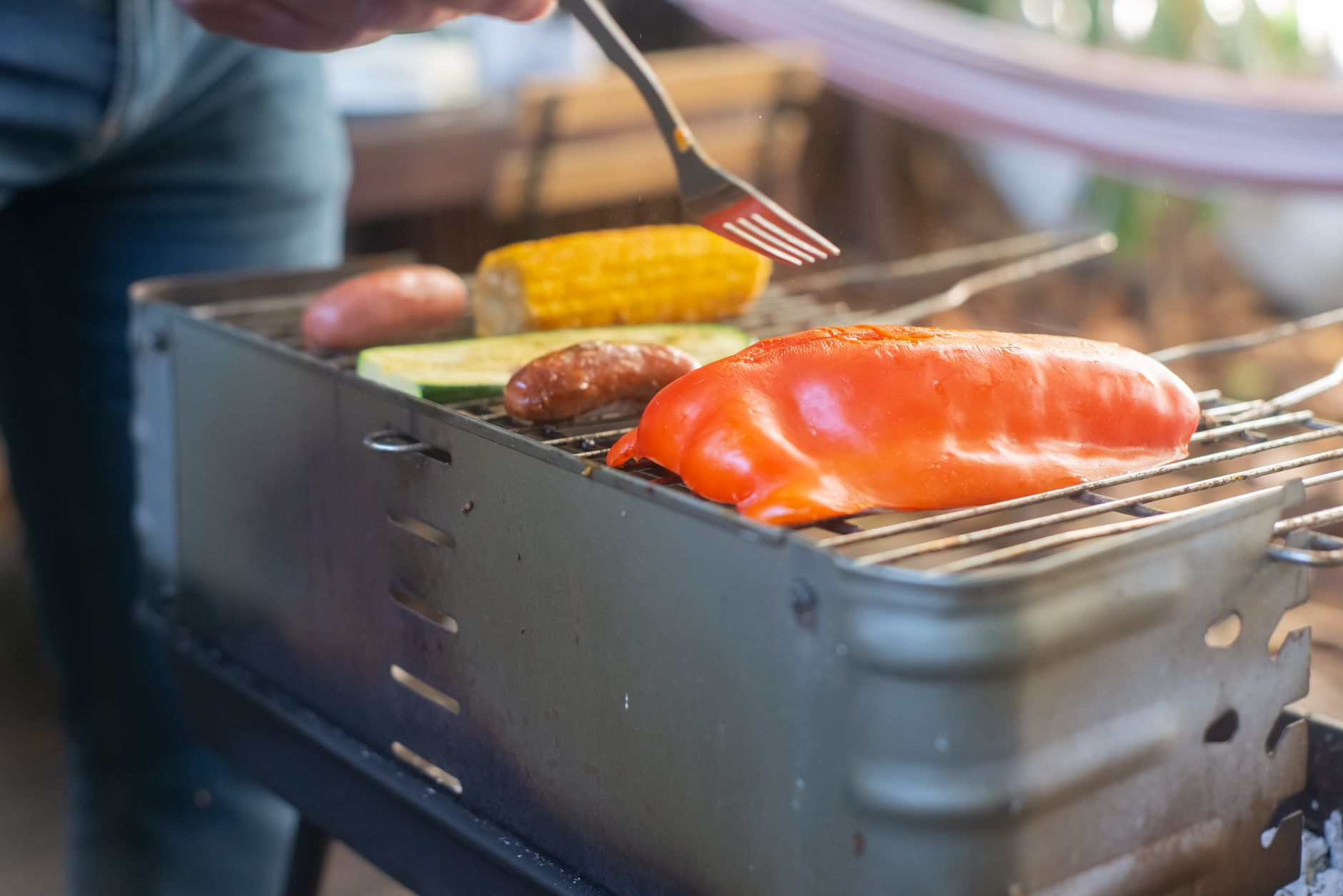 Close-up of bell pepper, corn, and sausages grilling on an outdoor barbecue.