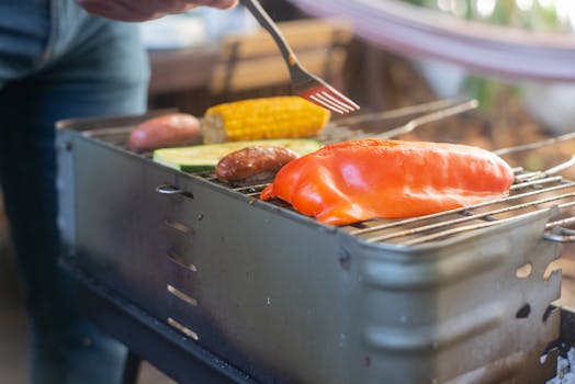 Close-up of bell pepper, corn, and sausages grilling on an outdoor barbecue.