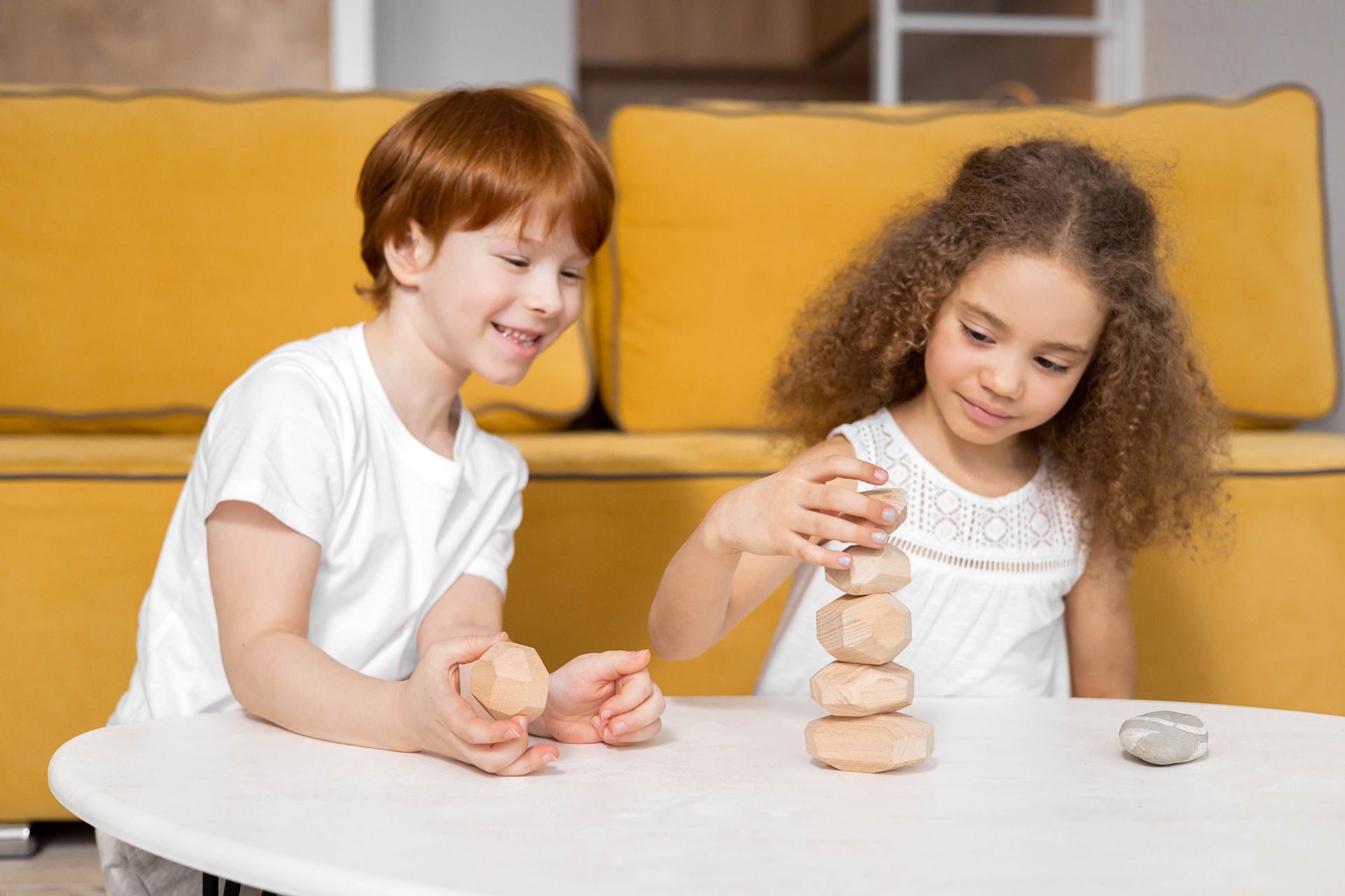 Two children playing with wooden stacking stones at a white table indoors.