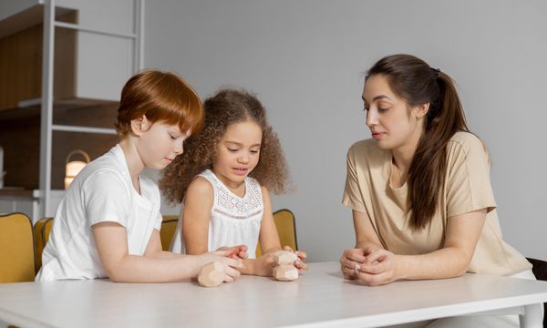 Babysitter — Children and woman sitting together at a white table, playing with wooden blocks.