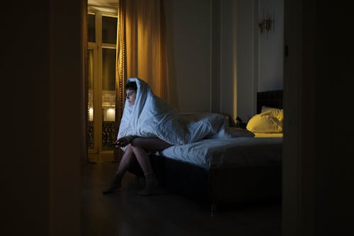 Photo of a Woman Sitting on the Bed while Covered by a White Blanket