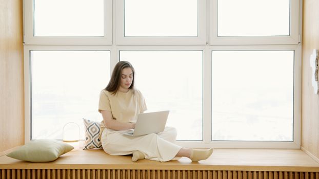 Woman sitting on windowsill working on laptop, creating a serene remote work environment.