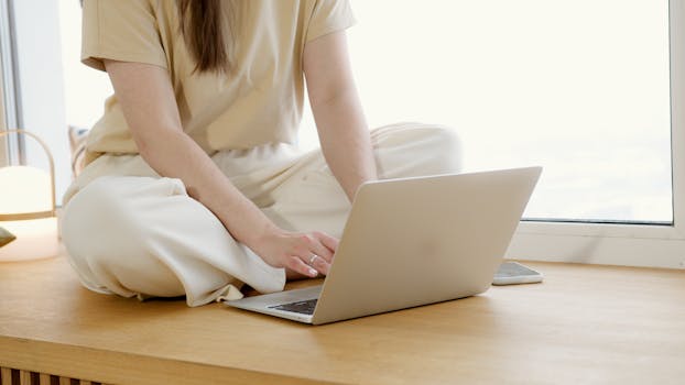 Unrecognizable woman sitting by a window, working on a laptop, embracing a relaxed work-from-home setup.