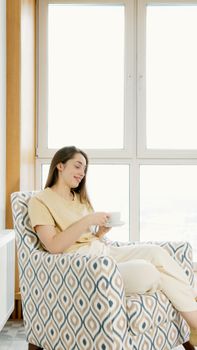 Serene woman enjoys coffee on a stylish chair by the window, embracing tranquility.
