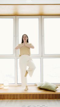 Caucasian woman in casual clothing practicing yoga indoors by a large window to enhance calm and balance.