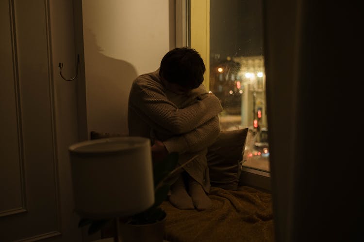 A Woman Sitting By A Window Hugging Her Knees 