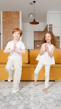 Two kids practicing yoga indoors, standing in tree pose with smiles, showcasing a sense of balance and enjoyment.