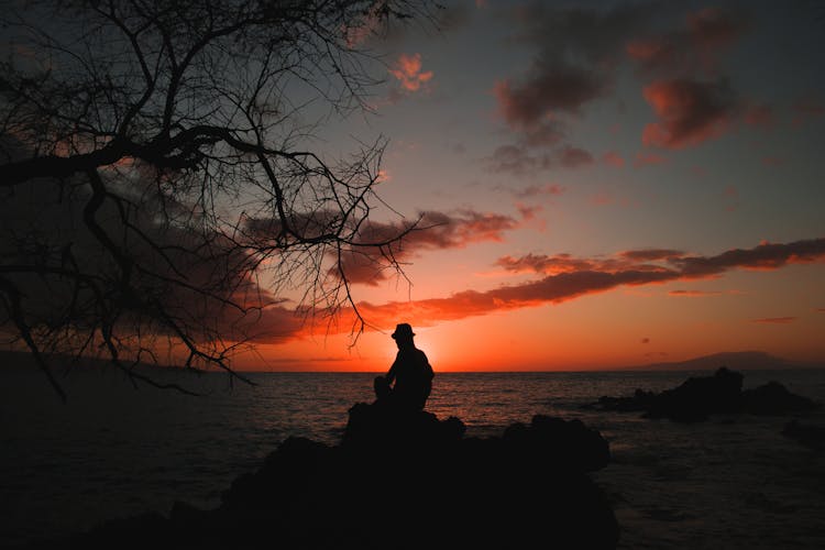 Silouhette Of Person Seating On Stones Behind Body Of Water