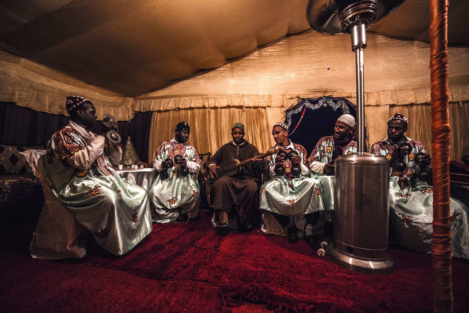 Group of African men in traditional clothes sitting on chairs placed on vivid red carpet and playing on national musical instruments in spacious tent