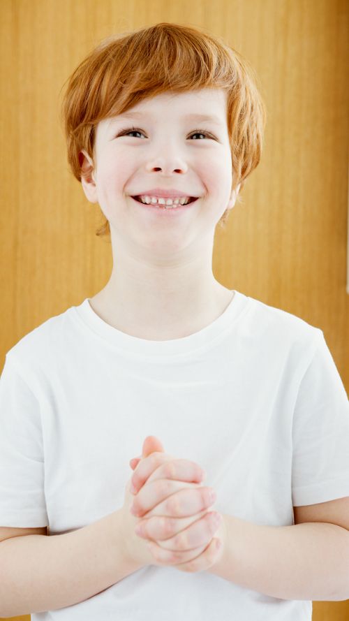 Free A cheerful child with red hair smiles warmly while clasping hands, wearing a white t-shirt indoors. Stock Photo