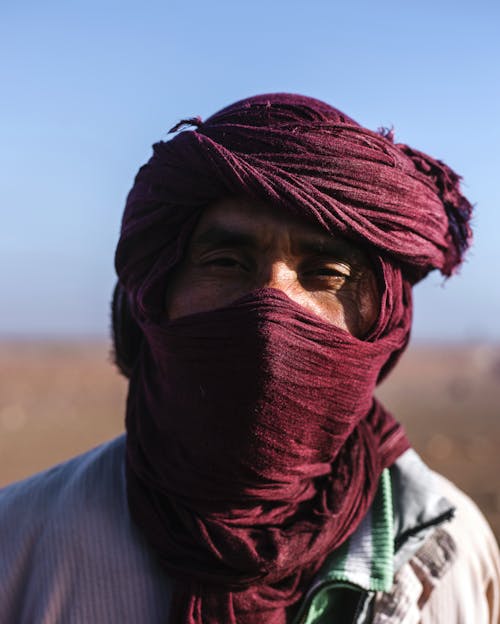 Free Portrait of unrecognizable ethnic man in traditional oriental headdress covering face with handkerchief while standing in desert and looking at camera on daytime Stock Photo