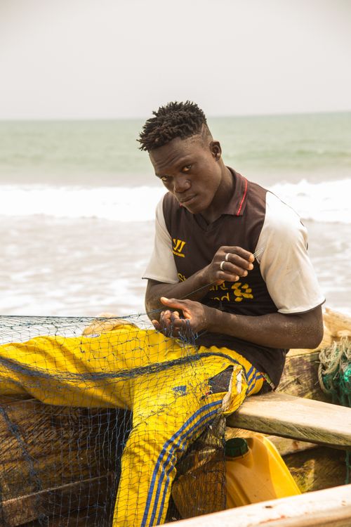 Free Portrait of a man sitting on a wooden boat by the sea, mending a fishing net in vibrant yellow pants. Stock Photo