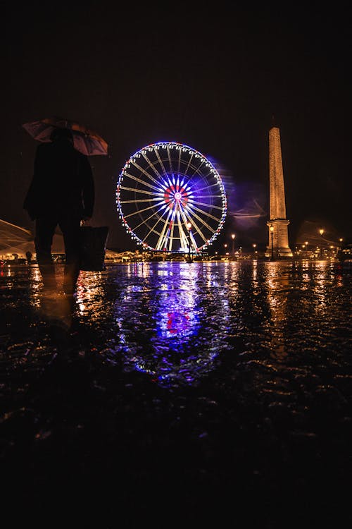Silhouette of faceless person with umbrella standing on wet square and admiring Ferris wheel with illumination located near column monument on night