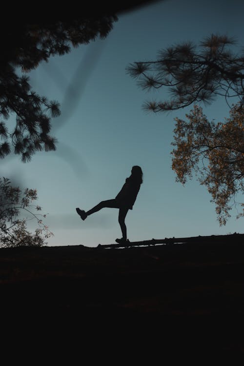 Silhouette of unrecognizable person standing with raised leg on top of hill in evening between branches of various trees