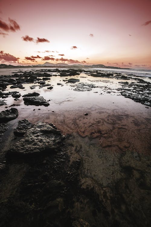 Drone view picturesque landscape of rough rocky coast covered with calm water against colorful sky with rare clouds during sundown