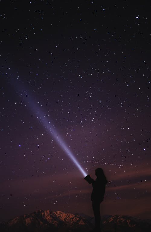 Side view silhouette of anonymous person standing on top of hill and sending ray from flashlight to dark night sky with stars