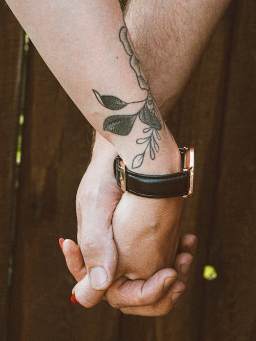 Crop anonymous loving young tattooed woman and man holding hands while standing against wooden fence in nature