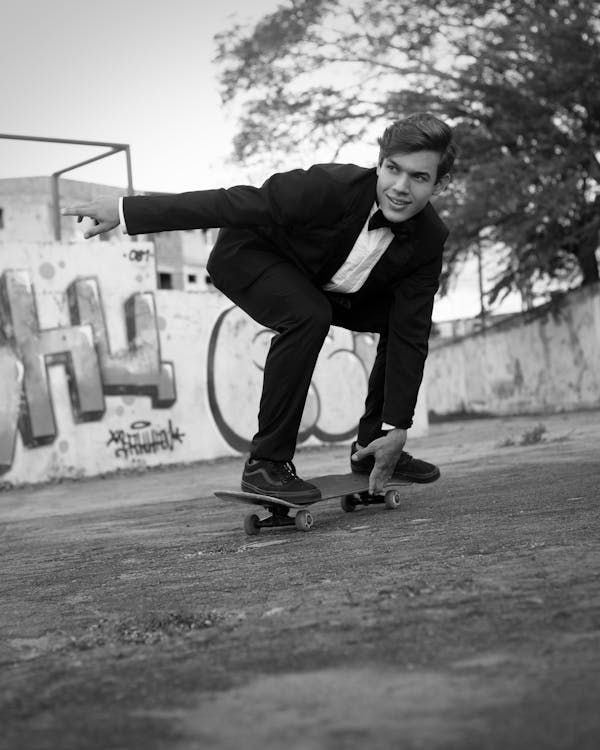 Black and white of positive young male millennial in elegant suit with bow tie smiling while riding skateboard in shabby park near graffiti walls