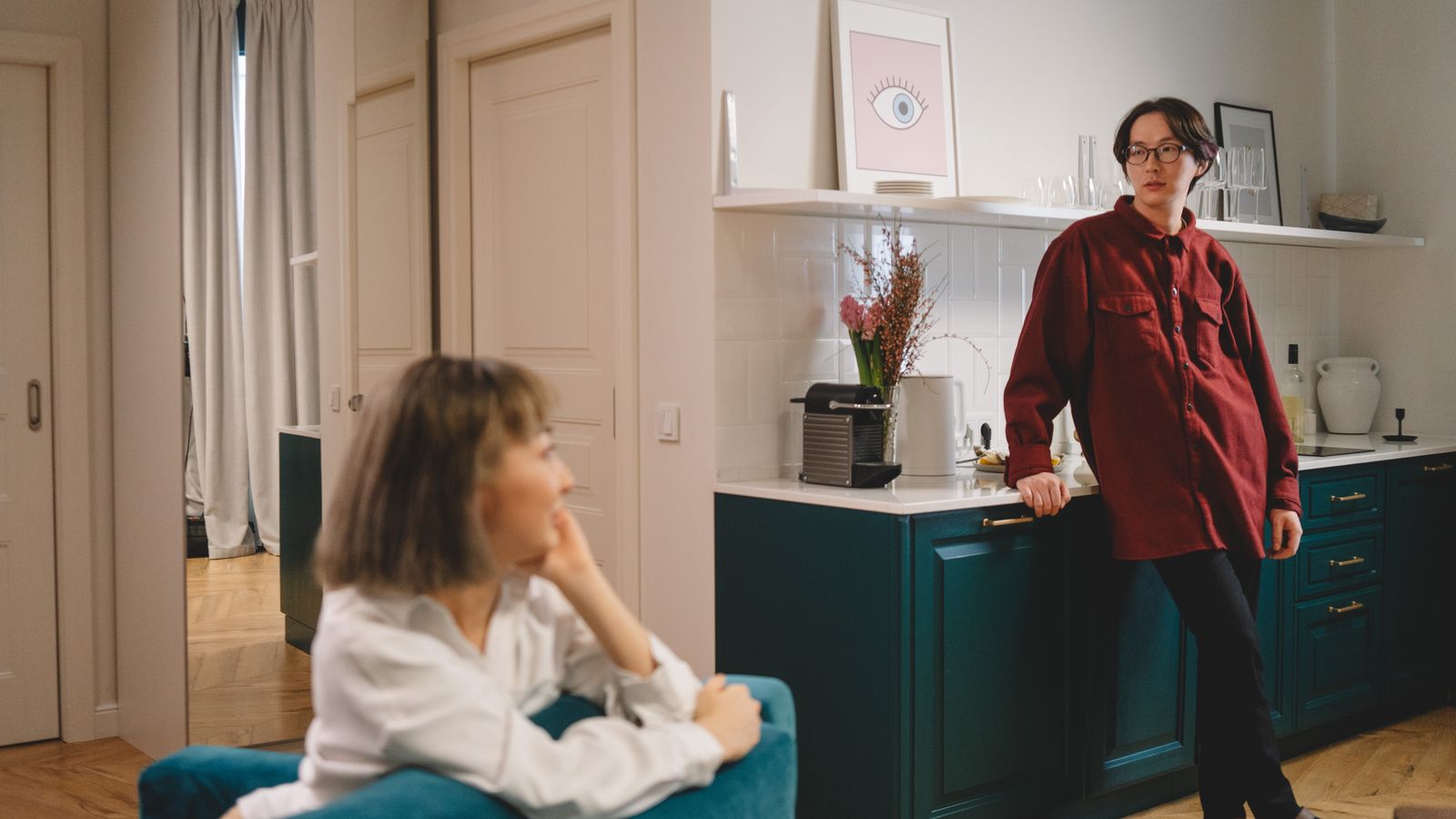 Two adults talking, one seated and one standing beside a kitchen counter