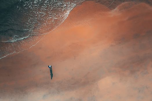 Aerial view of unrecognizable traveler relaxing on wide sandy beach near wavy ocean on sunny day