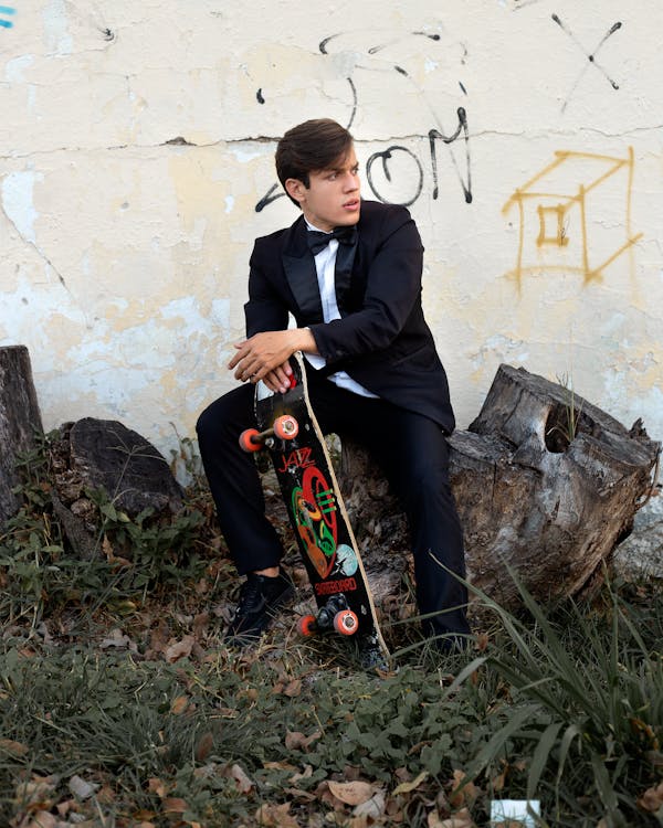 Stylish guy in classy suit sitting on log with skateboard in hand