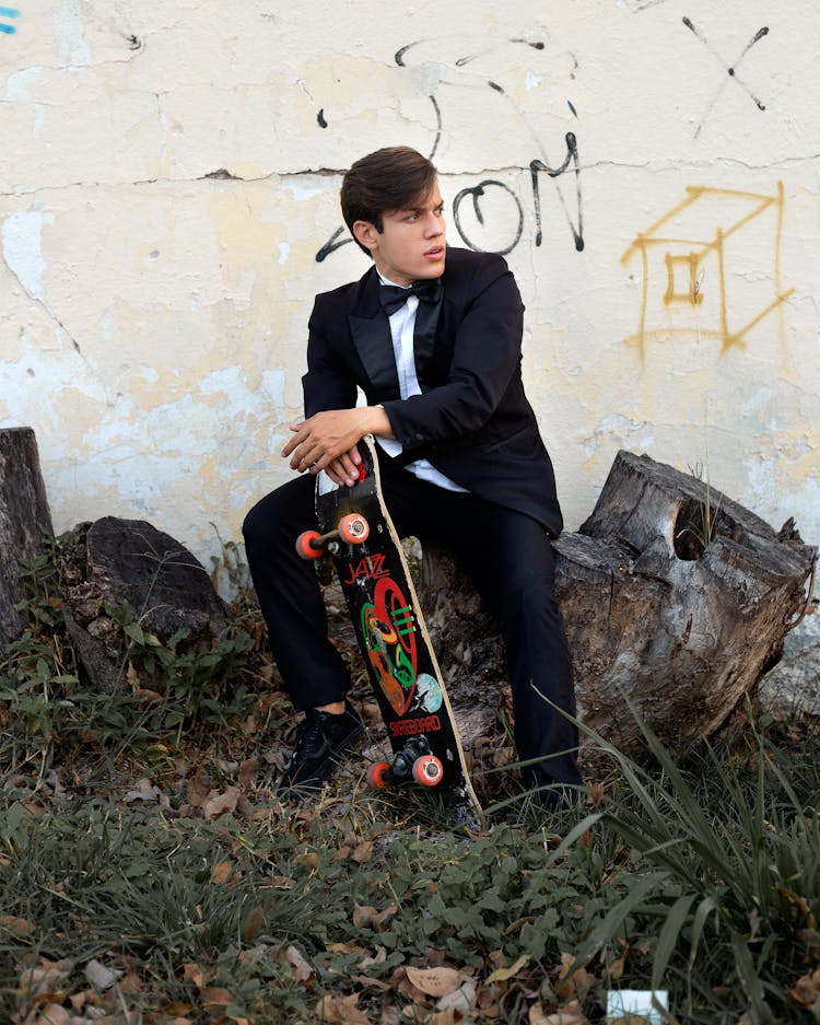 Stylish Guy In Classy Suit Sitting On Log With Skateboard In Hand