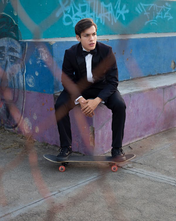 Full body of handsome young male millennial in classy suit and bow tie with skateboard sitting on shabby concrete stairs with graffiti and looking away