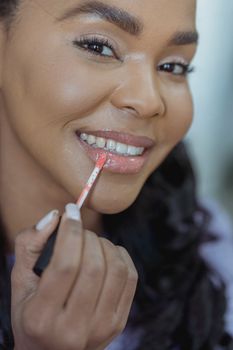 Crop positive African American female applying lip gloss while doing makeup and looking at camera