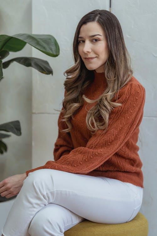 Charming female with long wavy hair sitting on seat near potted plant and looking at camera