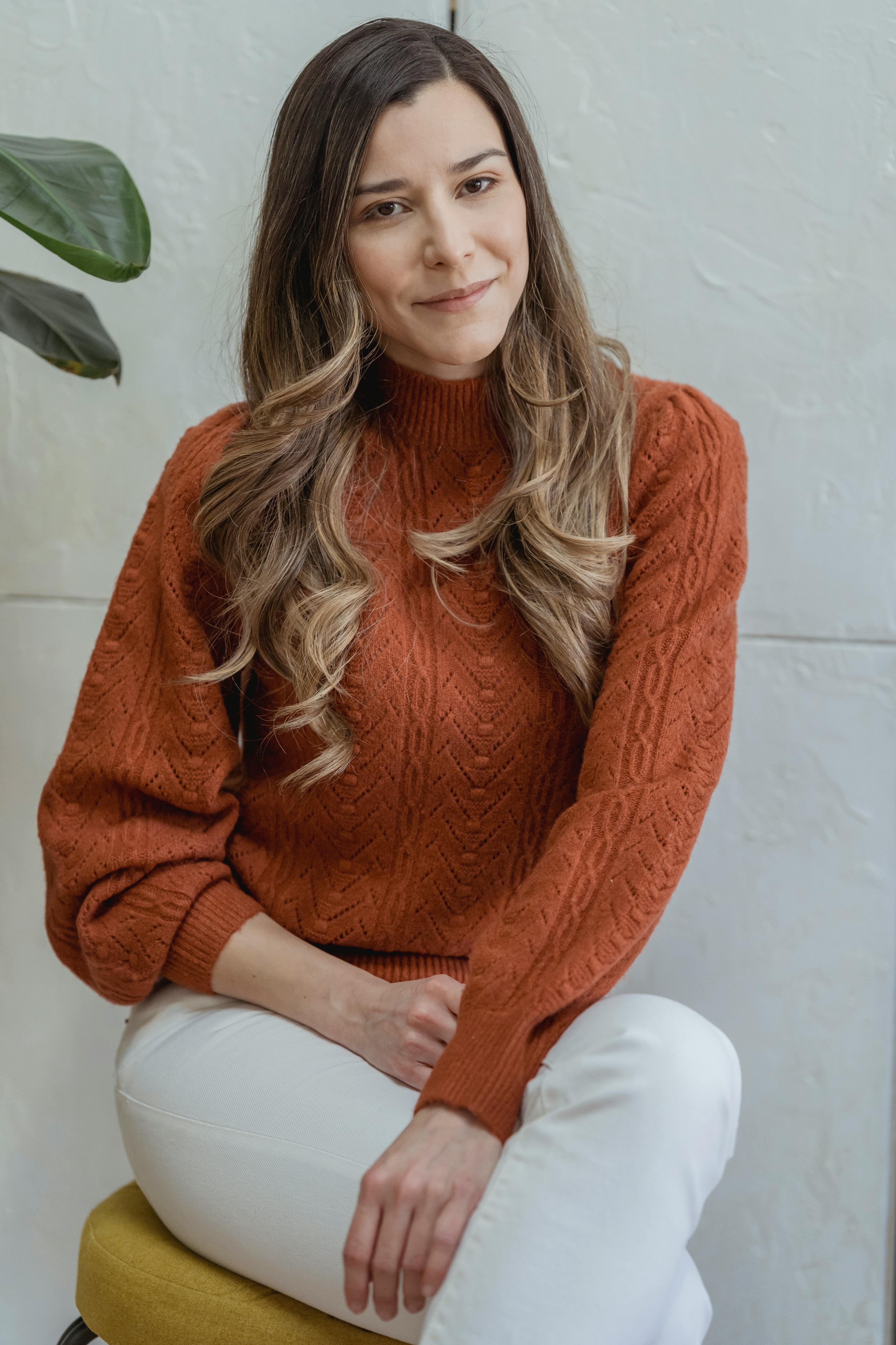 Charming female in sweater and skinny pants sitting on pouf with crossed legs and looking at camera