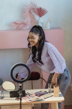 Side view of African American at table with eyeshadow palettes and makeup brushes in room with ring lamp while recording online lesson