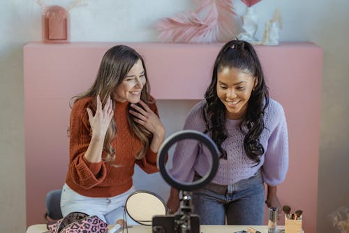 Free Positive multiethnic female makeup artists at table with professional supplies looking at smartphone near ring lamp while recording online lesson Stock Photo
