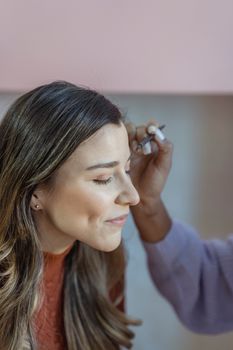 Unrecognizable female visagiste applying cosmetic product on eyebrow of cheerful female client with closed eyes during makeup procedure in beauty salon