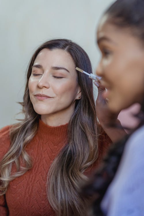 Free Side view of crop African American female makeup artist applying eyeshadow on eyelids of young woman sitting in salon with closed eyes Stock Photo