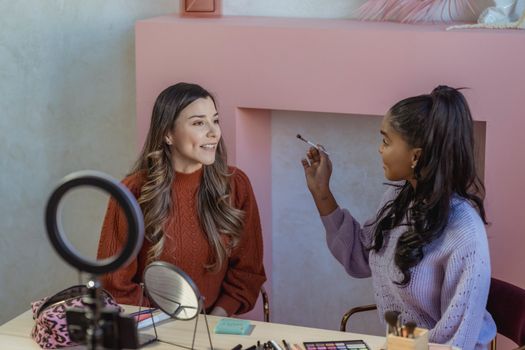Positive young African American lady in stylish sweater applying makeup on face of female friend sitting at table with mirrors and various cosmetic products