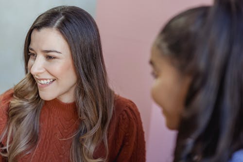 Cheerful young multiracial female best friends in casual clothes smiling while chatting with each other at home