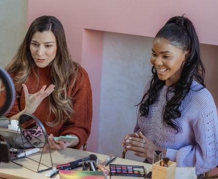 Diverse friends smiling and speaking online at table with round shaped mirror and eyeshadows with brushes