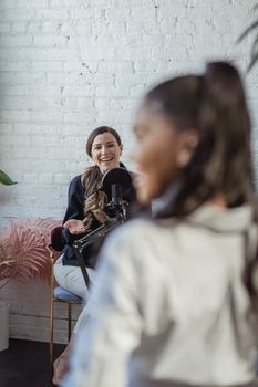 Diverse cheerful women smiling while recording voice with microphone in modern studio with white brick walls