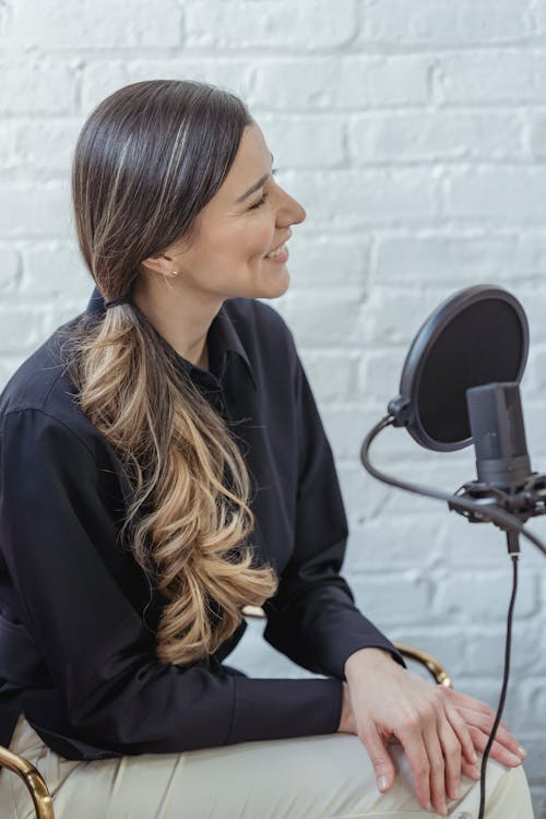Glad female with long wavy hair in black shirt talking while recording voice in modern studio with brick walls