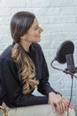 Glad female with long wavy hair in black shirt talking while recording voice in modern studio with brick walls